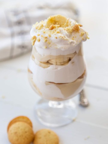 A glass with banana pudding that has layers of the pudding, whipped cream, banana slices, and vanilla wafers.