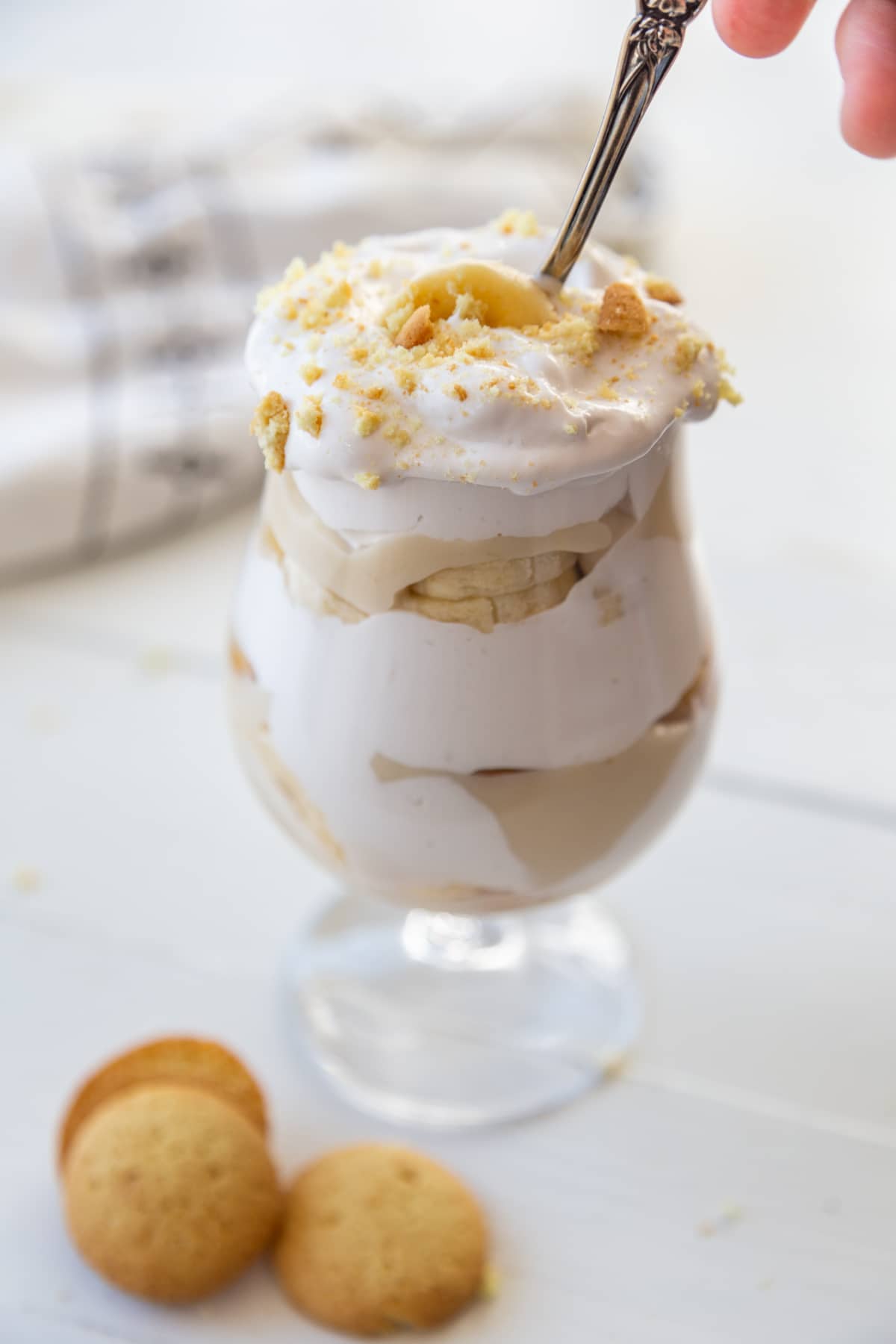A hand inserting a silver spoon into a glass of banana pudding with 3 vanilla wafers next to the glass. 