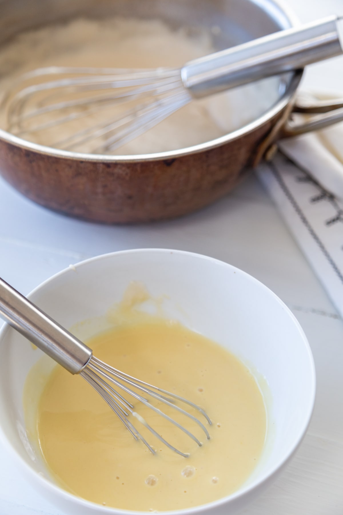A white bowl with egg and a whisk and a copper pot with vanilla pudding in the background. 
