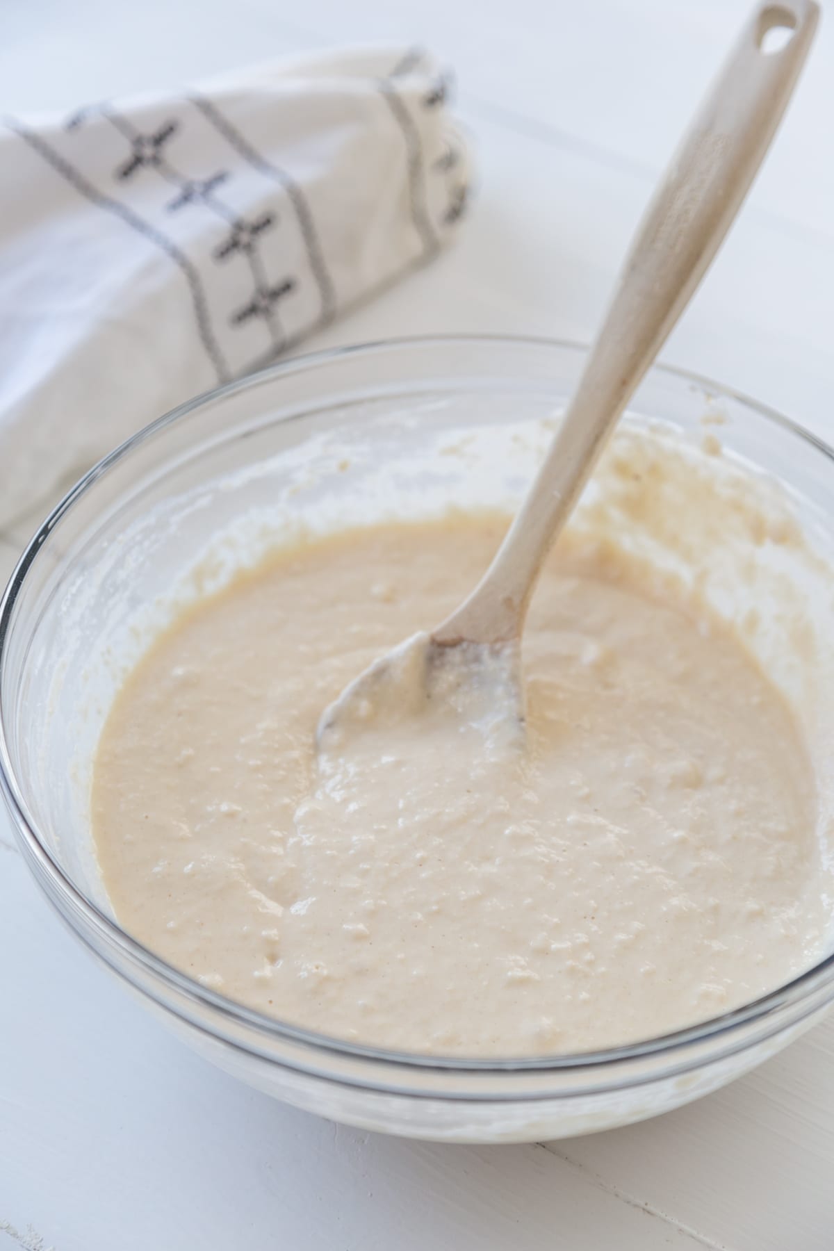 A glass bowl with waffle batter and a wooden spoon in the bowl and a white and black towel next to the bowl.