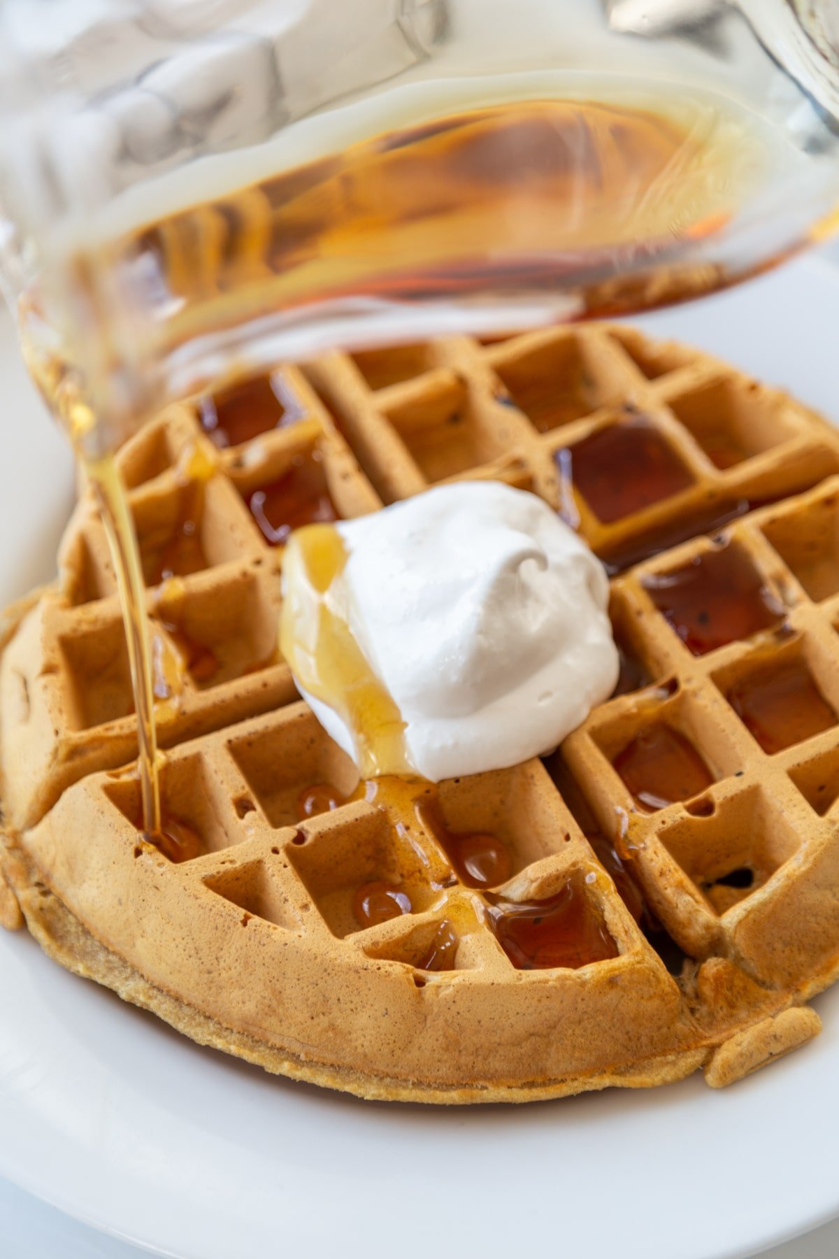 A Belgian waffle with maple syrup being poured over the waffle and whipped topping on a white plate. 