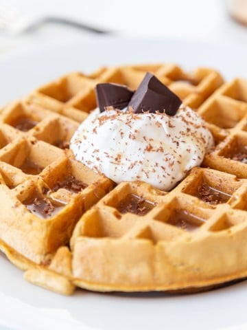 A Belgian waffle with whipped topping, chocolate shavings, and chocolate pieces sticking out of the whipped cream on a white plate.