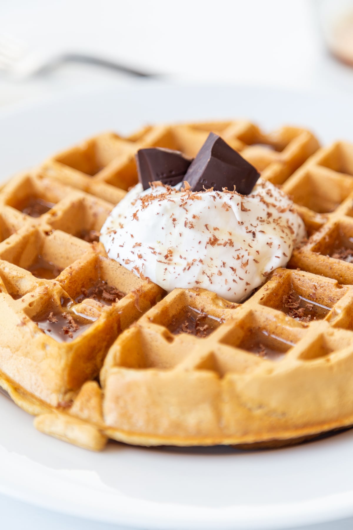A Belgian waffle with whipped topping, chocolate shavings, and chocolate pieces sticking out of the whipped cream on a white plate. 