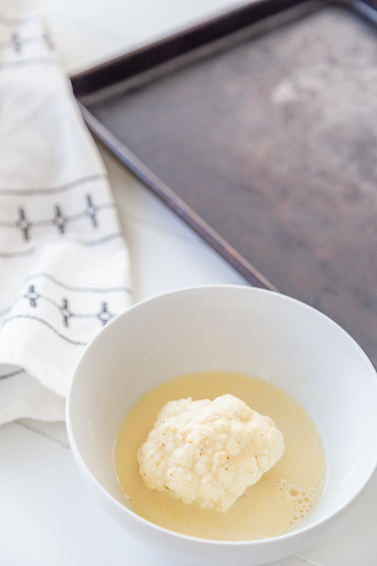 A white bowl with a cauliflower floret in an egg wash and a pan and white and black towel next to the bowl.