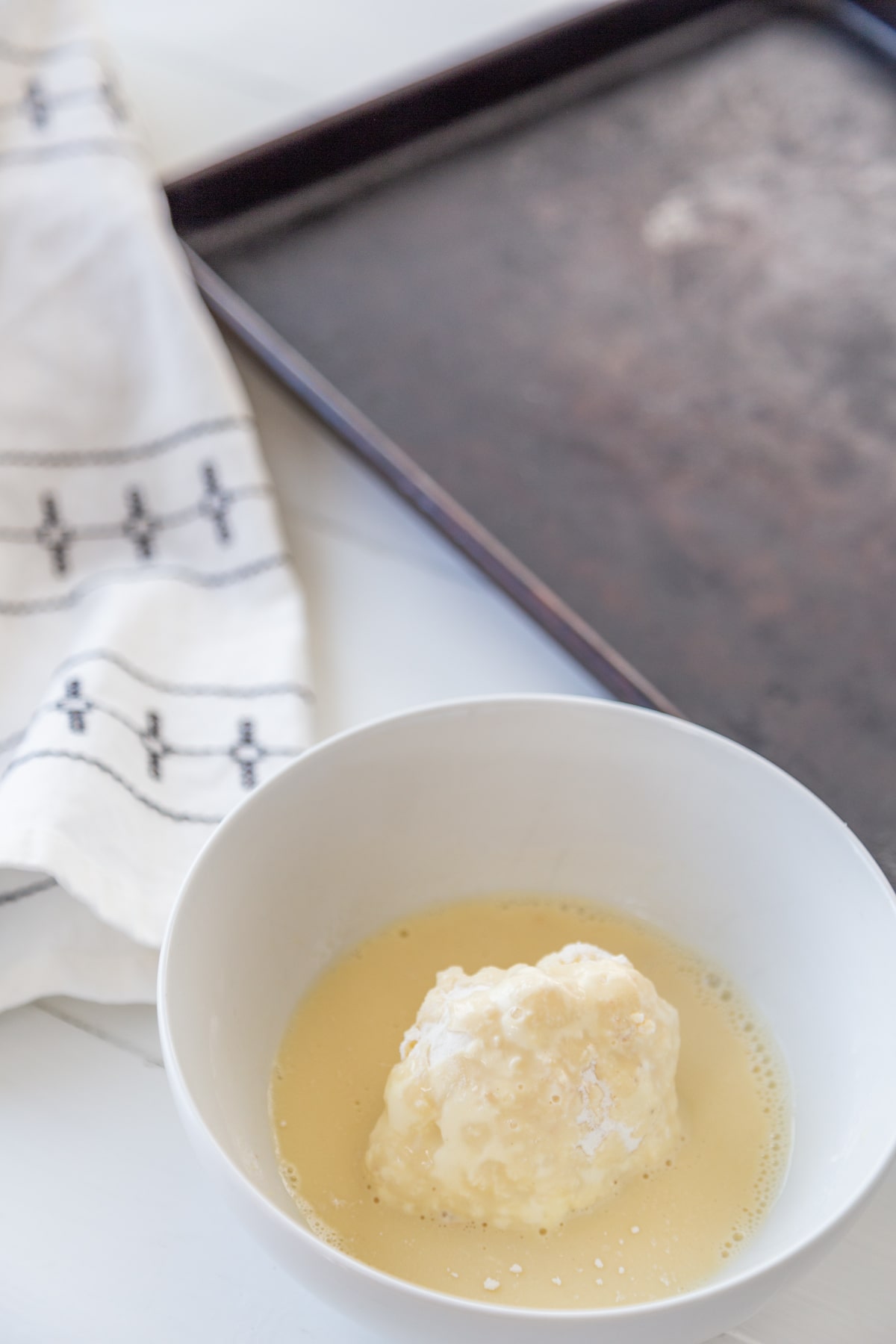 A cauliflower floret in a white bowl with egg and milk and a silver pan and white and black towel next to the bowl.