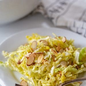 Shredded cabbage salad with slivered almonds on a white plate with a silver fork on the side of the plate and a white bowl of the salad in the background.