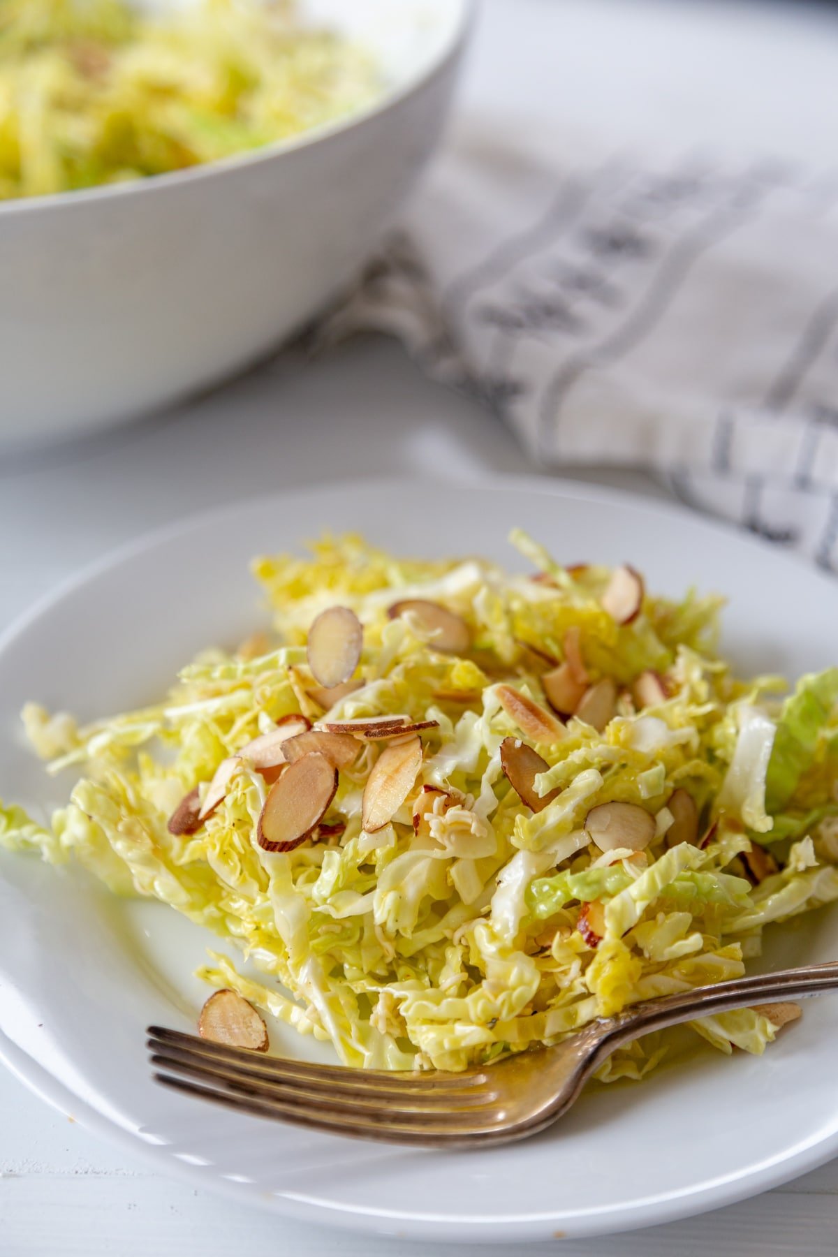 Shredded cabbage salad with slivered almonds on a white plate with a silver fork on the side of the plate and a white bowl of the salad in the background.