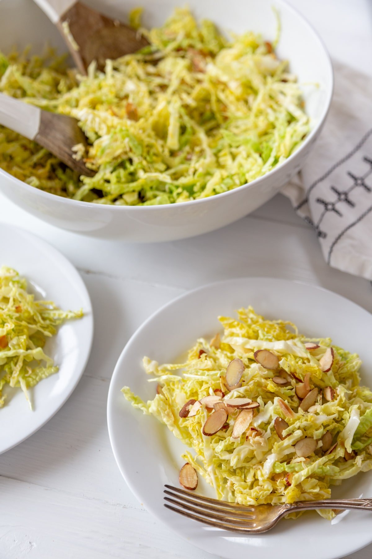 A white plate with shredded cabbage and almond salad with a silver fork on the edge of the plate, part of a white plate with salad on the left, and a white bowl of the salad with white and wood salad serving pieces in the bowl. 