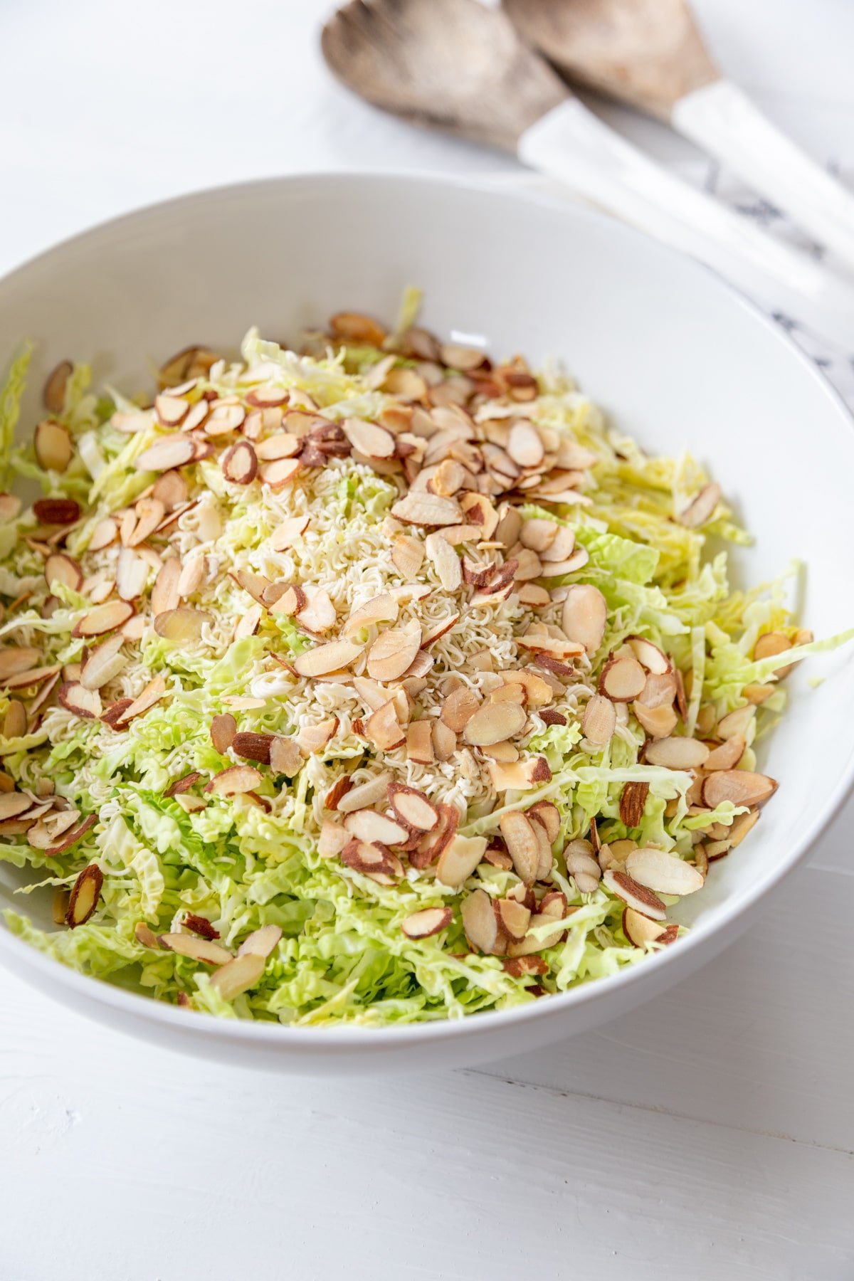 A white bowl with shredded green cabbage and toasted slivered almonds on top of it and a white and wood spoon and fork on the table next to the bowl. 