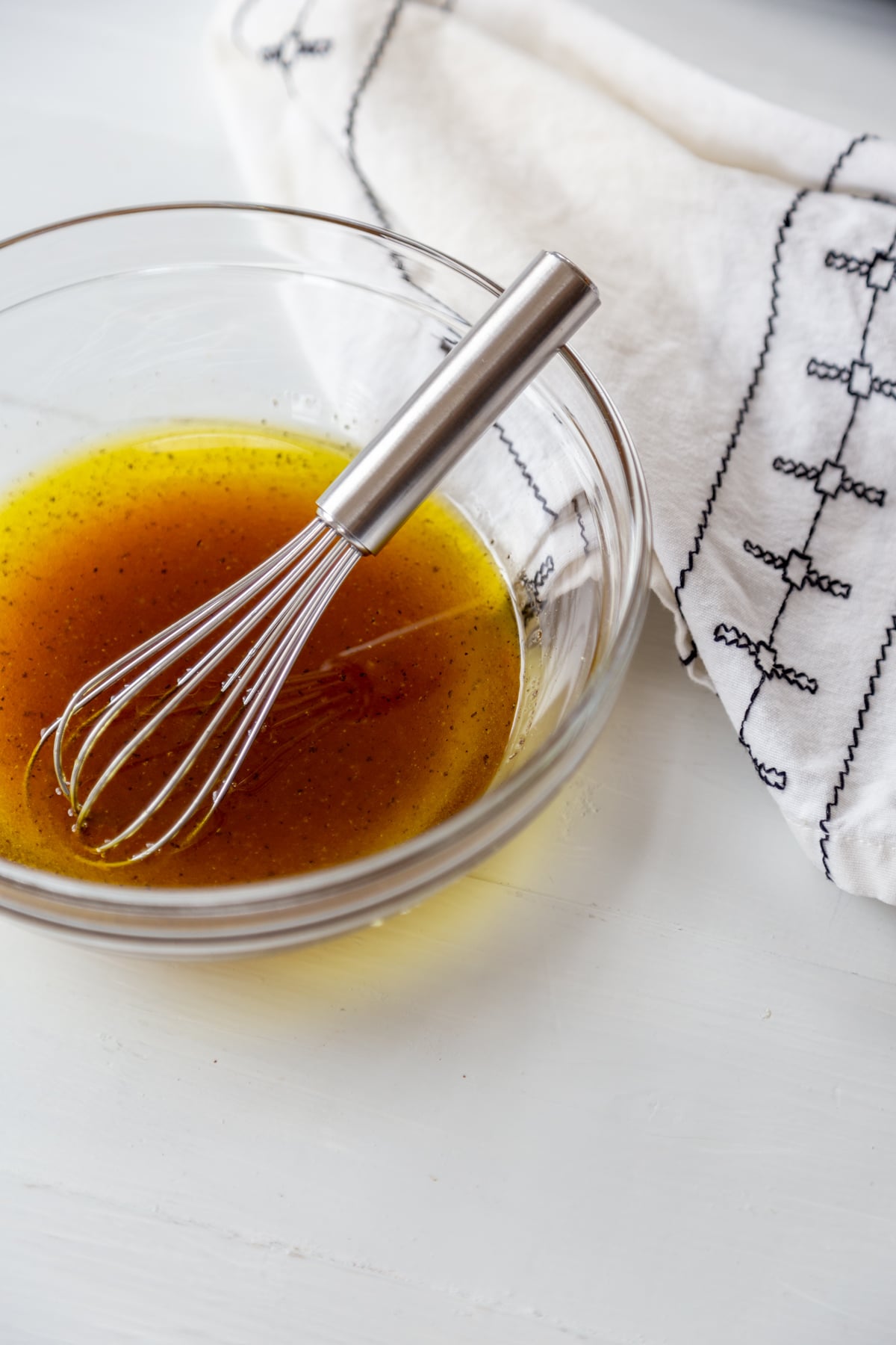 A clear glass bowl with soy dressing and a small silver whisk in the bowl, and a white and black towel next to the bowl. 