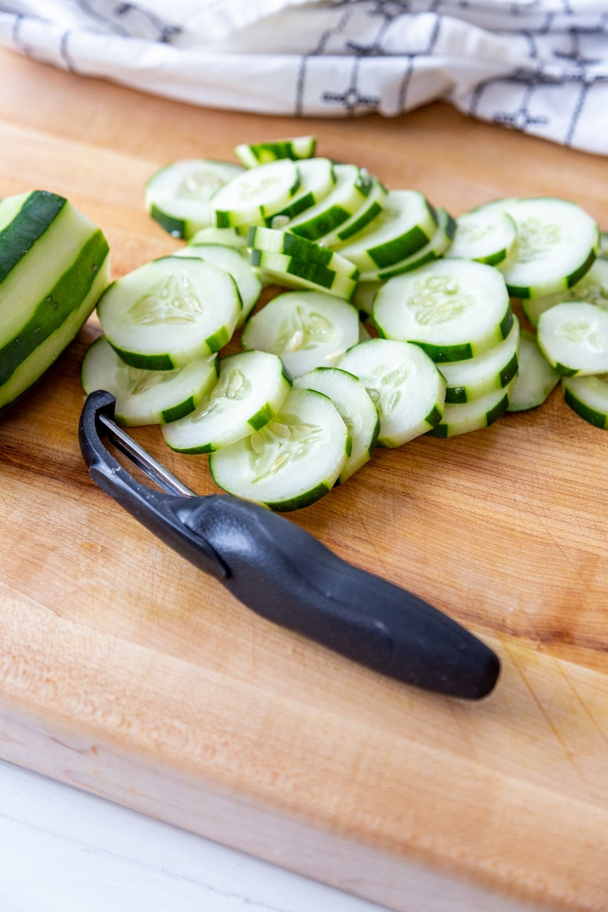 Sliced cucumbers on a wooden cutting board with a black vegetable peeler and a cucumber with stripes of skin peeled from it. 