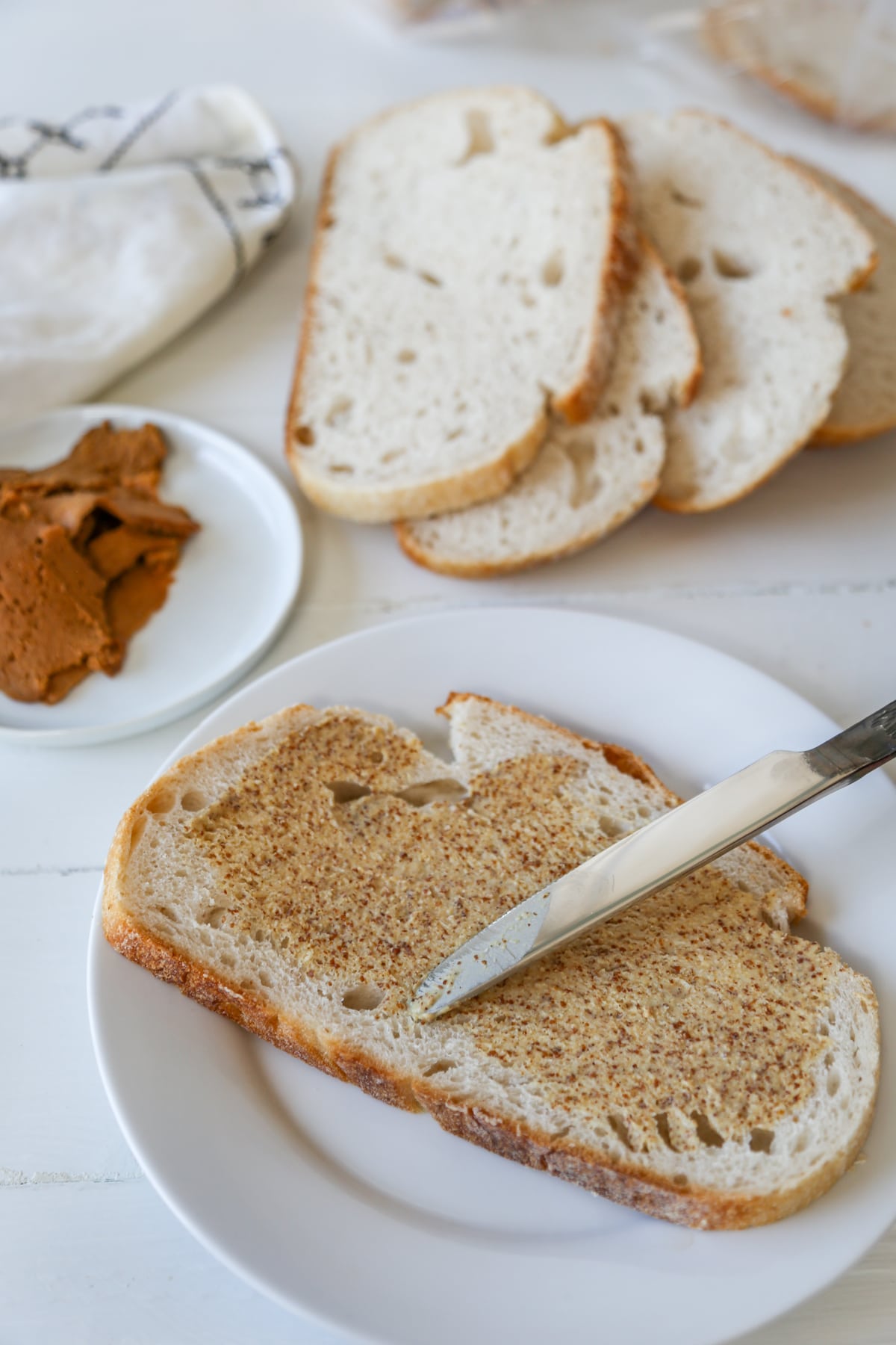 A knife spreading mustard on a slice of bread on a white plate with more bread slices and sliced ham in the background.