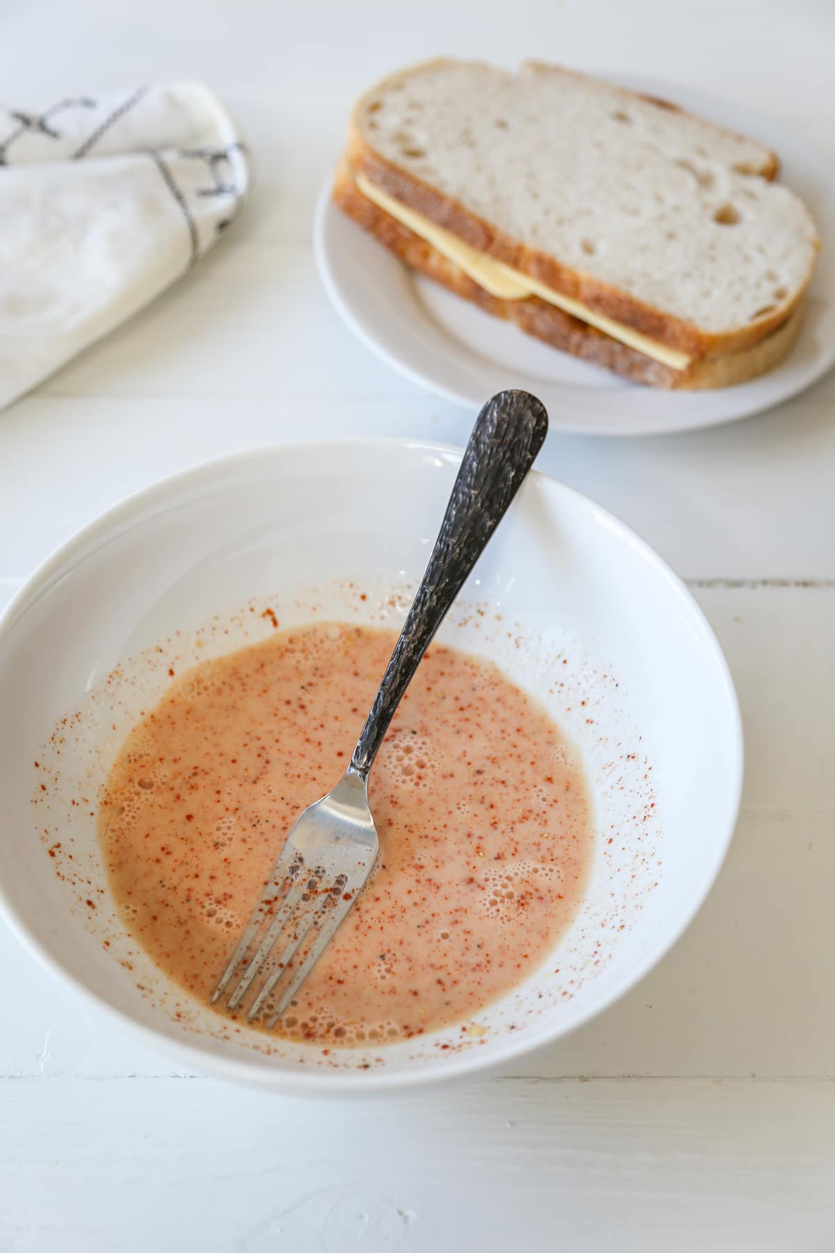 A white bowl with an egg wash mixed with orange spices and a fork in the bowl, and a ham and cheese sandwich next to it.