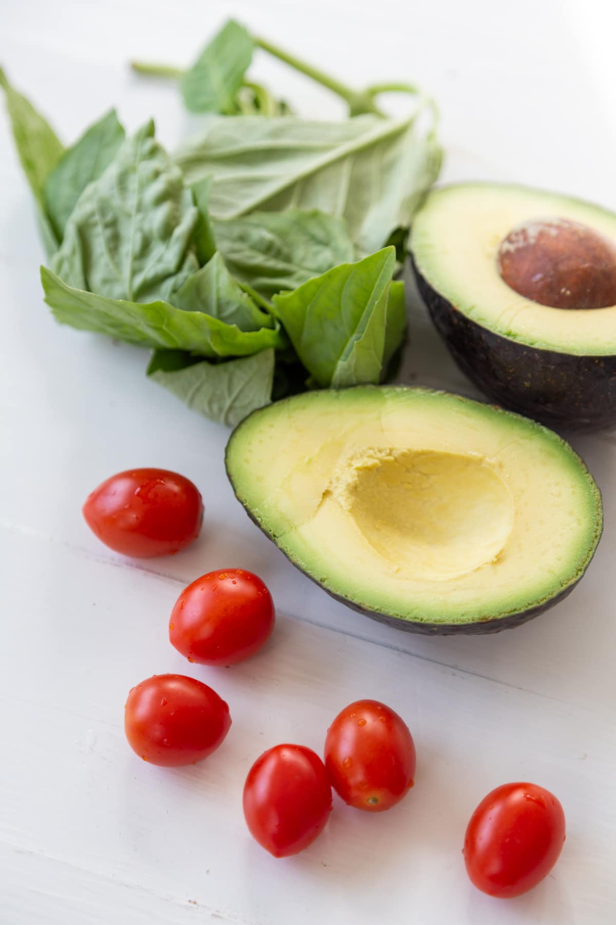 An avocado cut in half and 6 red cherry tomatoes and a bunch of basil leaves on a white surface.