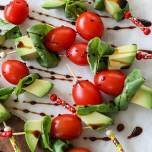 A marble and wood board with tomato, basil, and avocado skewers that are drizzled with balsamic vinegar.