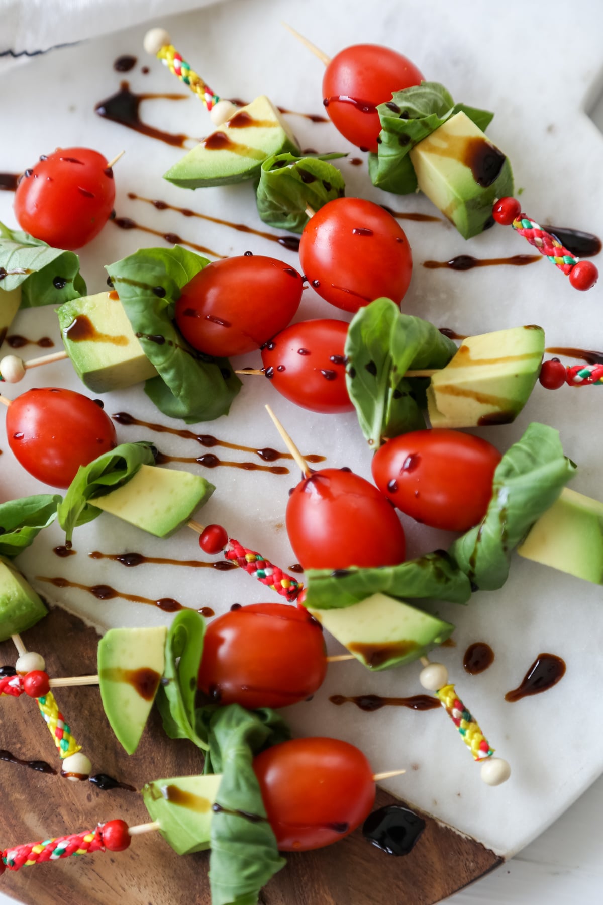 A marble and wood board with tomato, basil, and avocado skewers that are drizzled with balsamic vinegar. 