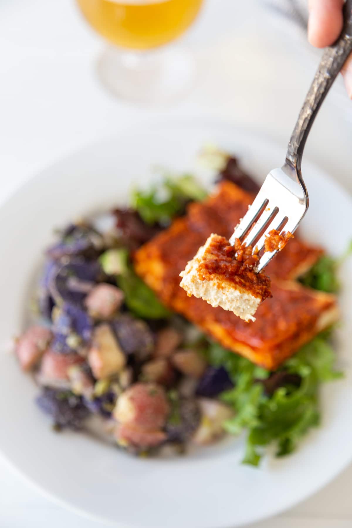 A silver fork with a piece of BBQ tofu held over a white plate with the tofu and potato salad.