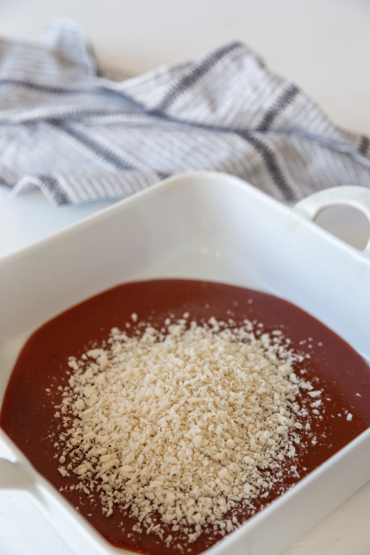 A square white baking dish with barbecue sauce and a pile of breadcrumbs in the center of the sauce.