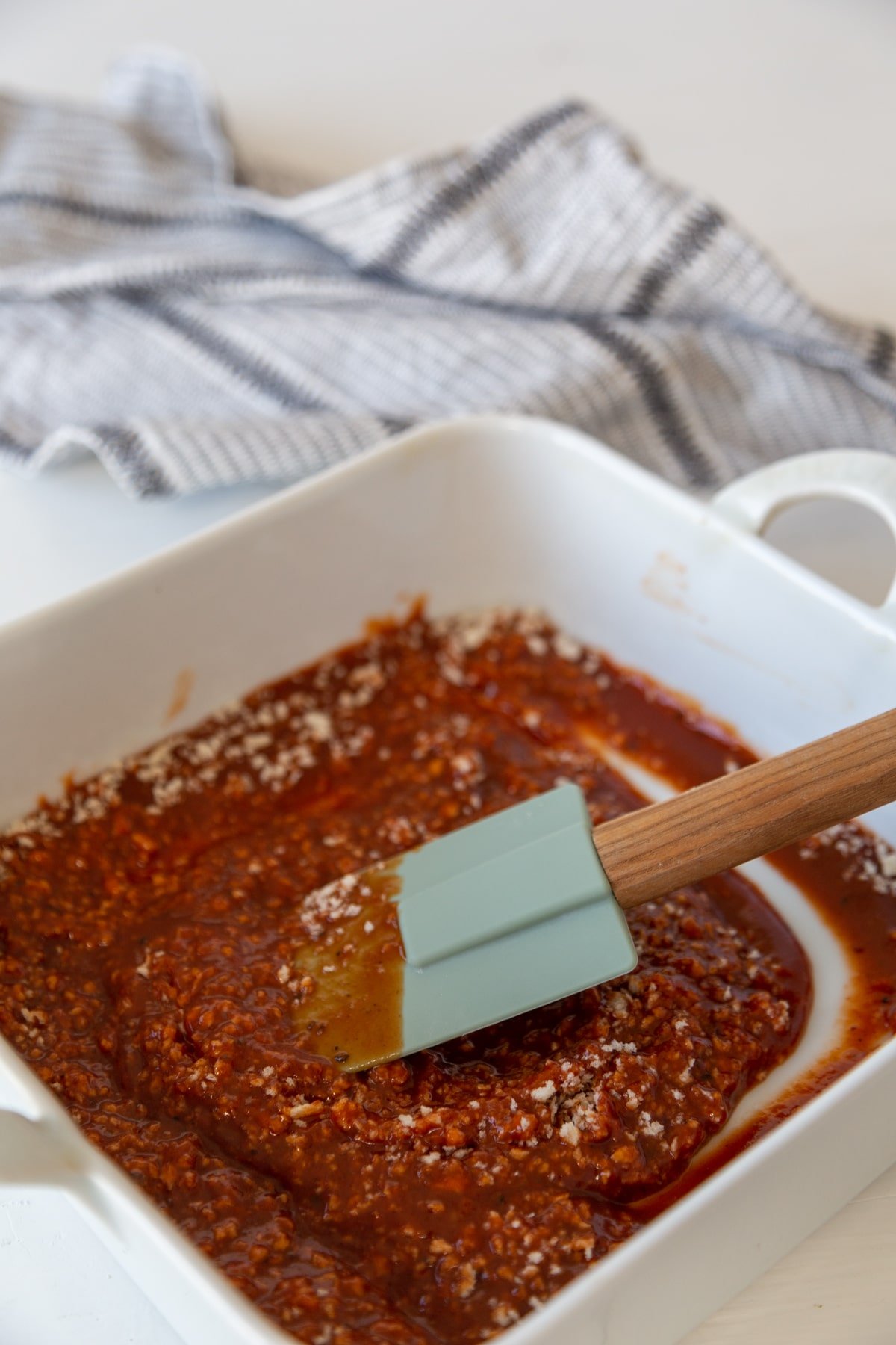 A green spatula stirring breadcrumbs into BBQ sauce in a square white baking dish.