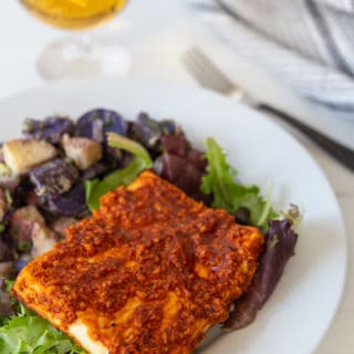 A piece of BBQ tofu on a white plate with purple and white potato salad, and a glass of beer in the background.