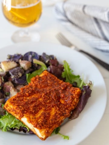 A piece of BBQ tofu on a white plate with purple and white potato salad, and a glass of beer in the background.