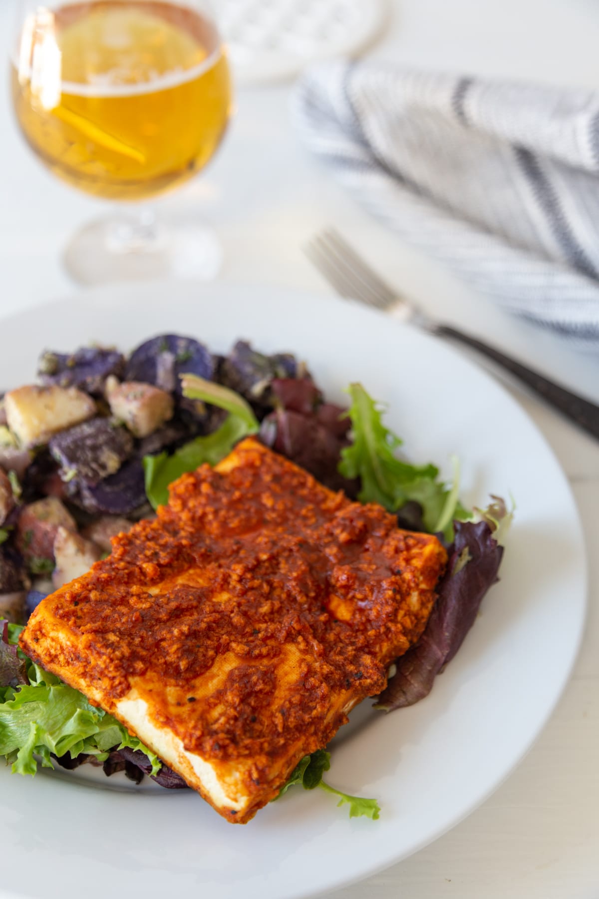 A piece of BBQ tofu on a white plate with purple and white potato salad, and a glass of beer in the background. 