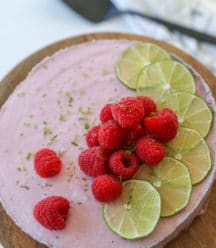 A raspberry cheesecake decorated with lime slices and whole raspberries on a wooden cake stand.