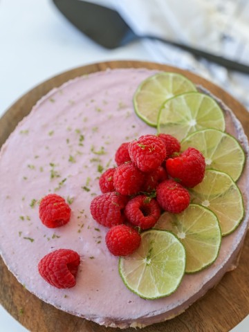 A raspberry cheesecake decorated with lime slices and whole raspberries on a wooden cake stand.