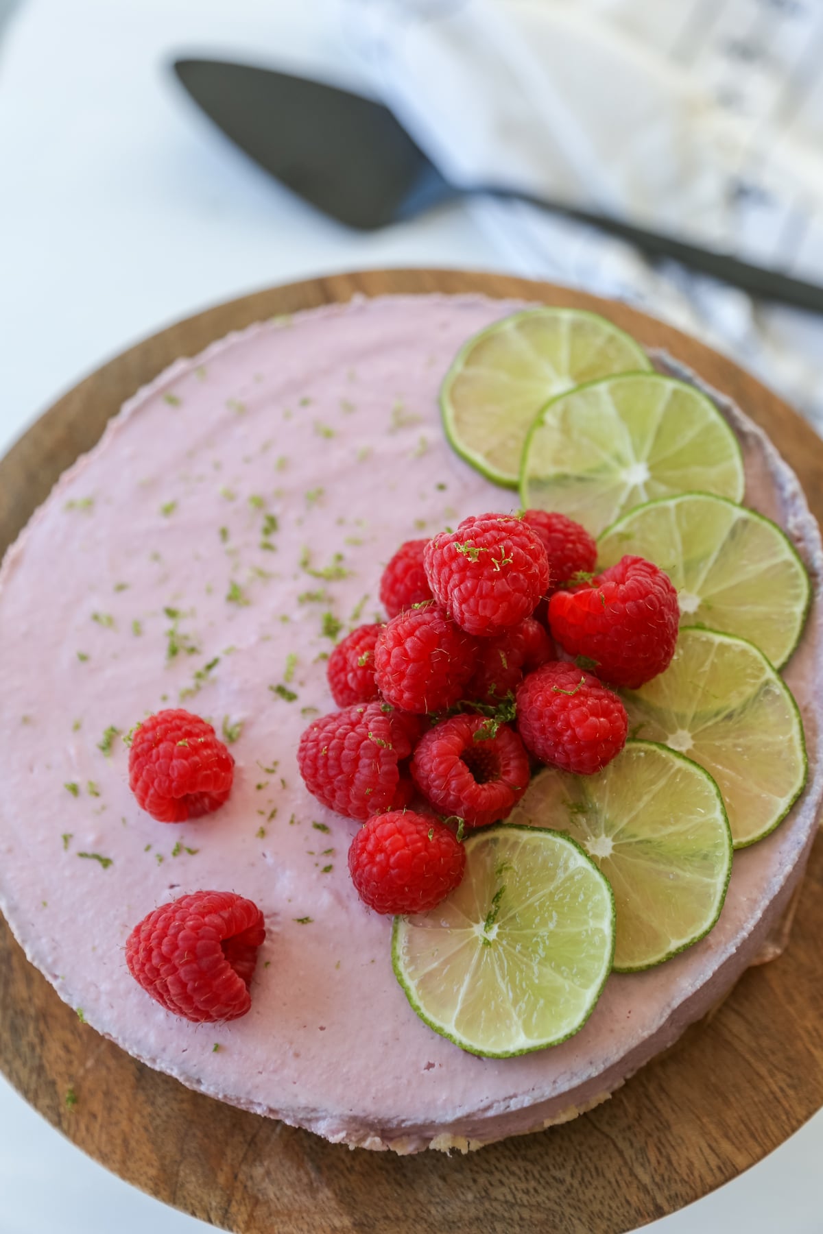 A raspberry cheesecake decorated with lime slices and whole raspberries on a wooden cake stand.