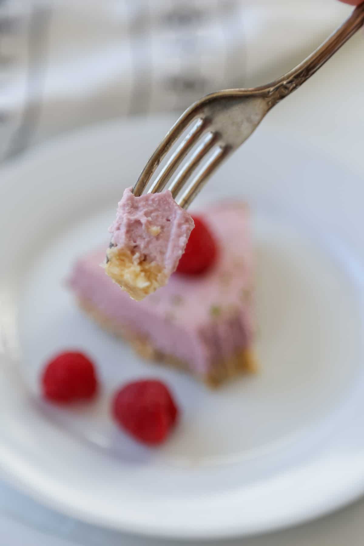 A silver fork with a piece of raspberry cheesecake held over a slice of the cake on a white plate with whole raspberries.