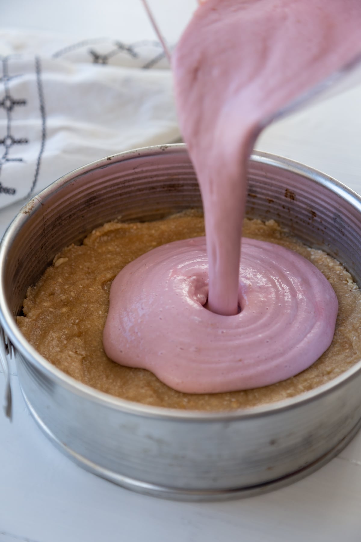 Raspberry cheesecake filling being poured into a springform pan with a nut crust on the bottom.