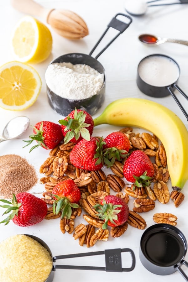 An ingredient photo for strawberry pecan waffles with measuring cups of flour, cornmeal, sugar, and strawberries, pecans, a banana, and lemon on a white table.