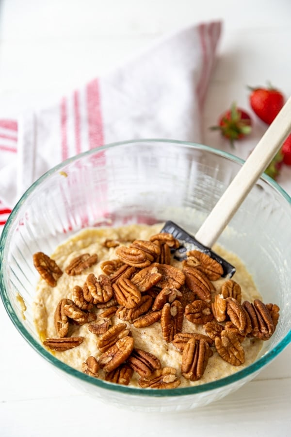 A glass mixing bowl with waffle batter and a spatula stirring toasted pecans into the batter.