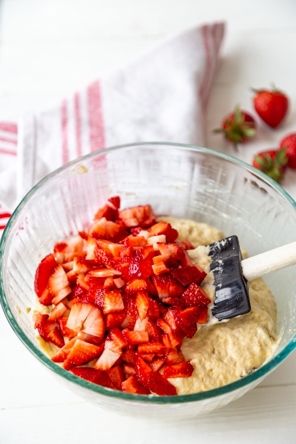 A glass mixing bowl with waffle batter and a spatula stirring strawberries into the batter.