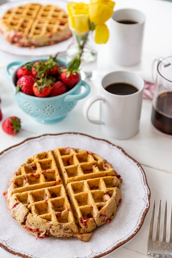 Two white plates with strawberry pecan Belgian waffles and a blue bowl of strawberries and 2 cups of coffee.