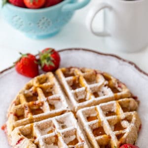A strawberry pecan Belgian waffle on a white plate with strawberries on the side and powdered sugar sprinkled on top, and a blue bowl of strawberries and a cup of coffee in the background.