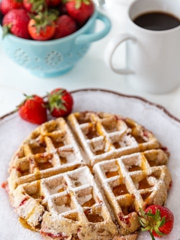 A strawberry pecan Belgian waffle on a white plate with strawberries on the side and powdered sugar sprinkled on top, and a blue bowl of strawberries and a cup of coffee in the background.