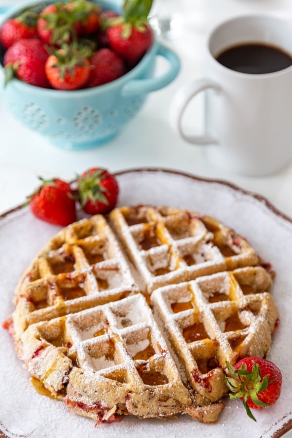 A strawberry pecan Belgian waffle on a white plate with strawberries on the side and powdered sugar sprinkled on top, and a blue bowl of strawberries and a cup of coffee in the background.