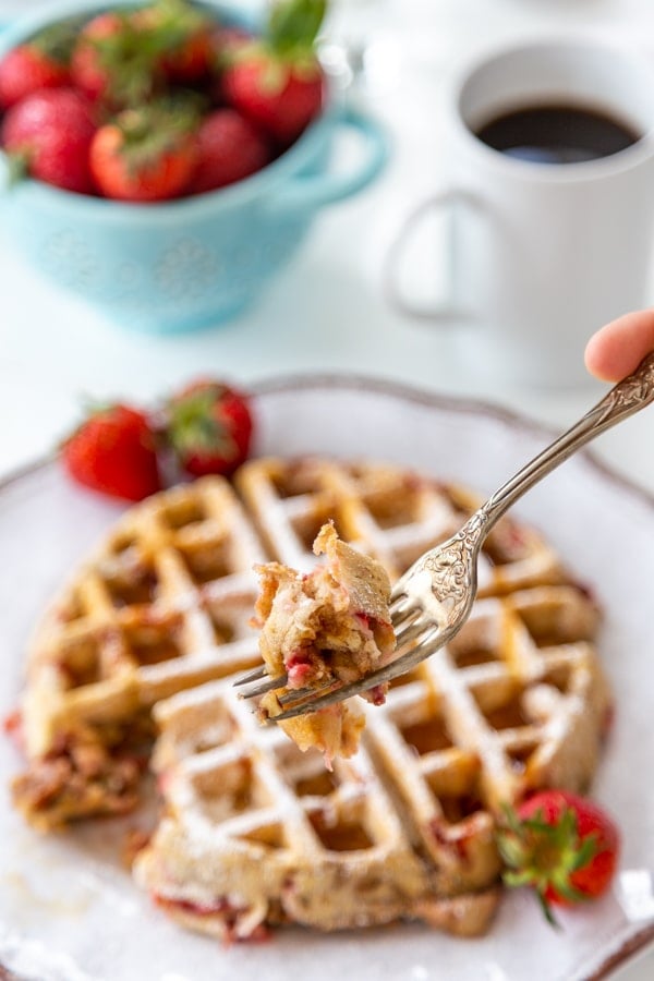 A fork full of a strawberry pecan waffle being held over a white plate with the waffle and a bowl of strawberries and cup of coffee.