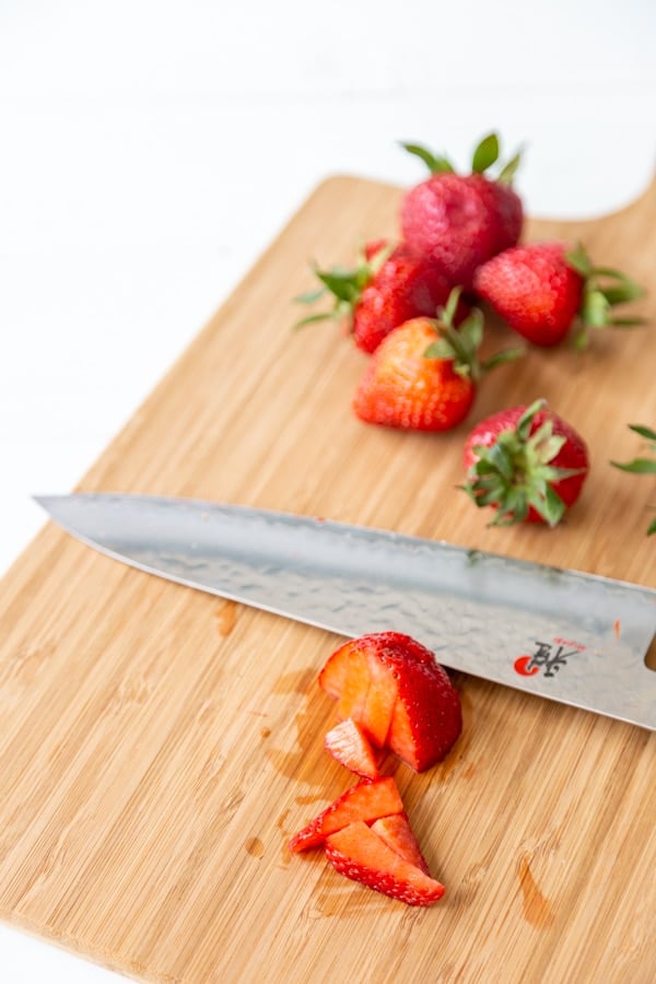 A wooden board with whole strawberries and a knife with sliced berries. 