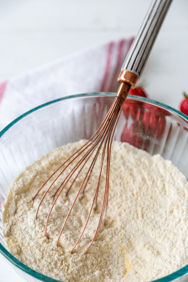 A glass mixing bowl with a copper whisk, whisking flour and cornmeal together.