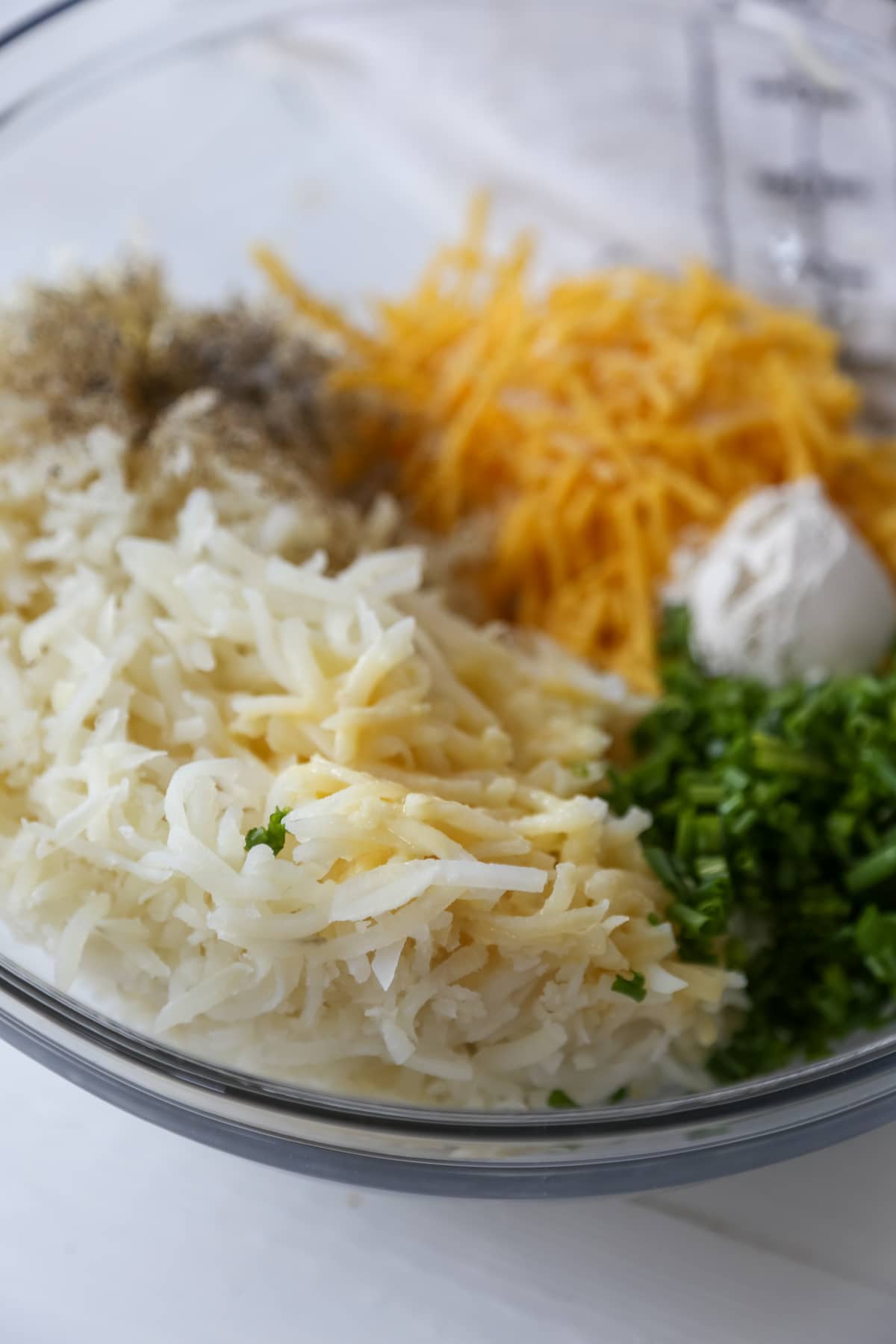 A glass bowl with shredded potato, shredded cheese, minced chives, and flour.