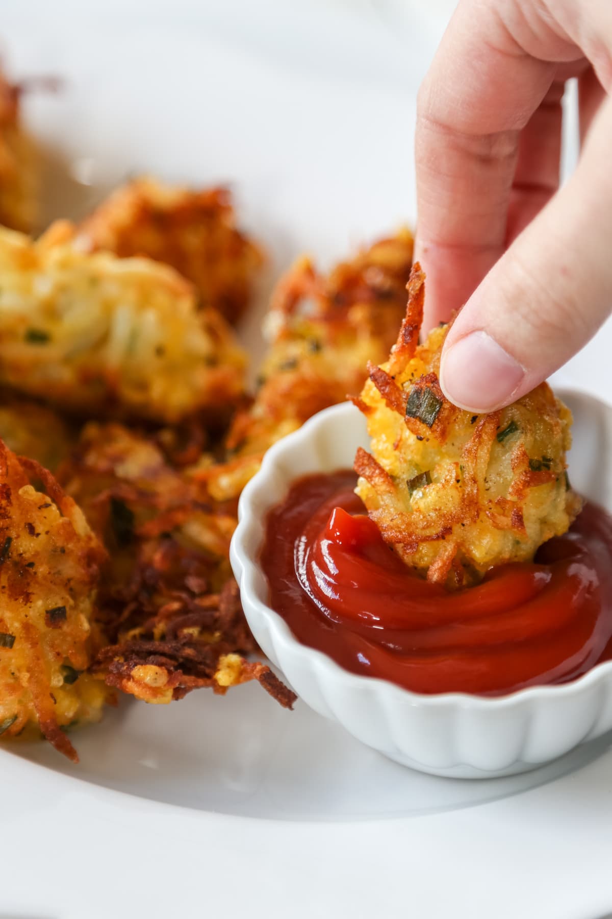 A hand dipping a tater tot into a bowl of ketchup that's on a white platter filled with tots.
