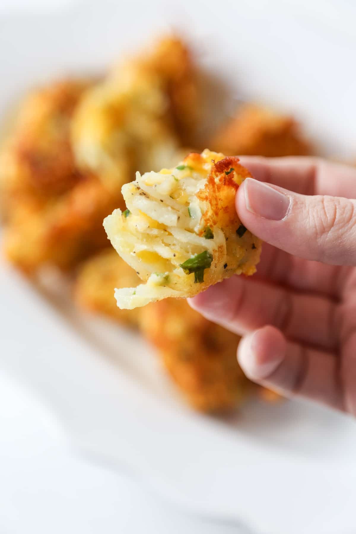 A hand holding half of a tater tot over a white platter of tots.