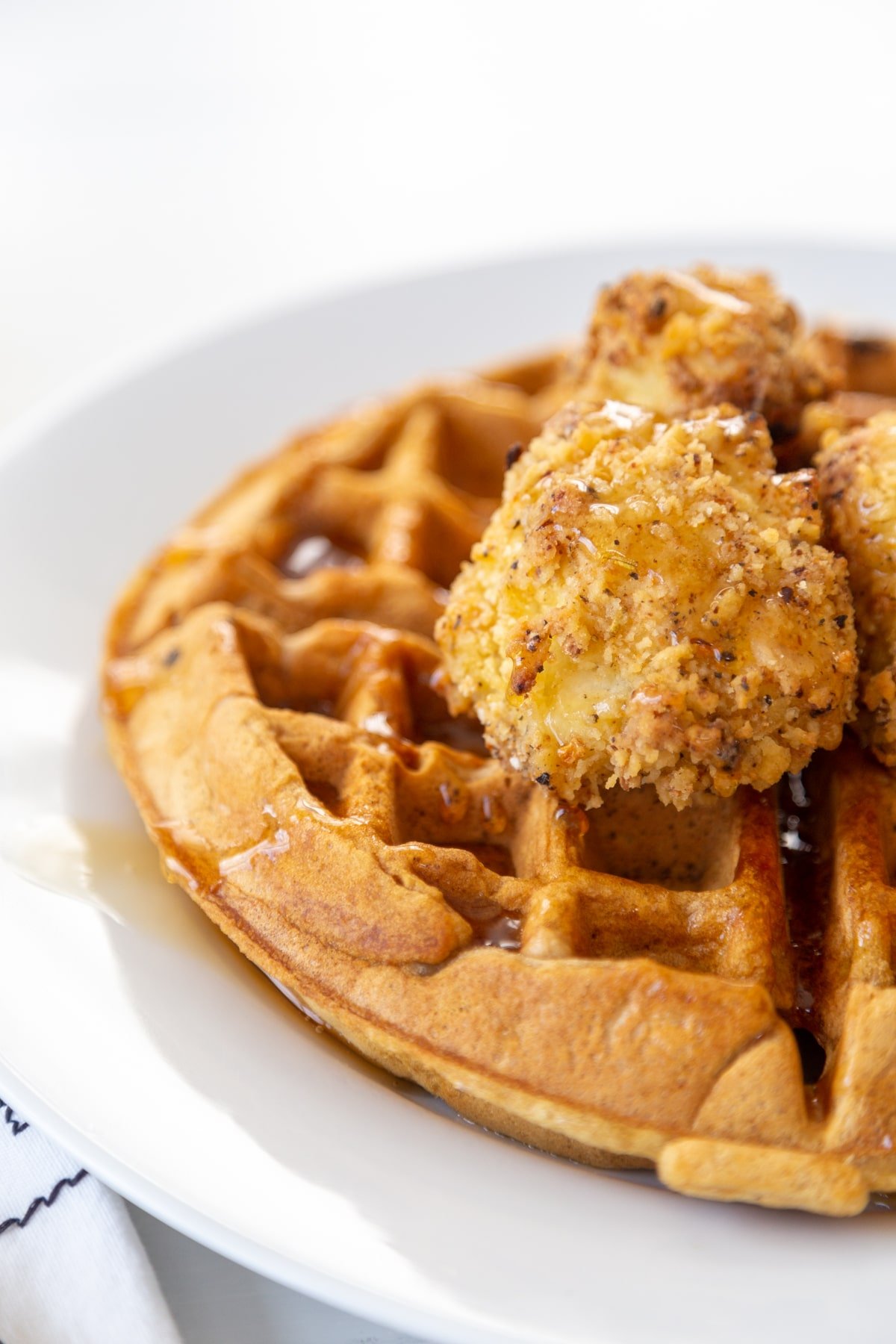 Half of a white plate with a Belgian waffle, crispy cauliflower on top and maple syrup drizzled over all of it.