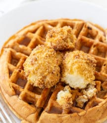 A Belgian waffle on a white plate with crispy cauliflower and maple syrup on top. A silver fork is next to the plate.