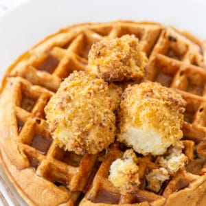 A Belgian waffle on a white plate with crispy cauliflower and maple syrup on top. A silver fork is next to the plate.