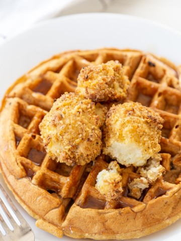 A Belgian waffle on a white plate with crispy cauliflower and maple syrup on top. A silver fork is next to the plate.