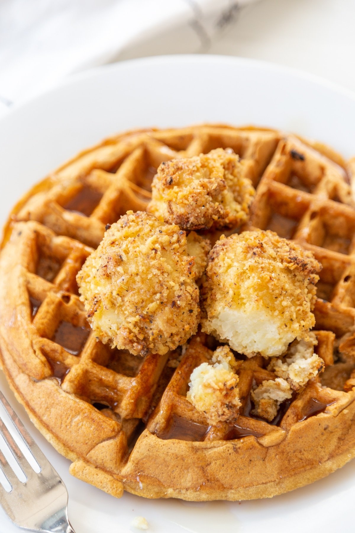 A Belgian waffle on a white plate with crispy cauliflower and maple syrup on top. A silver fork is next to the plate.