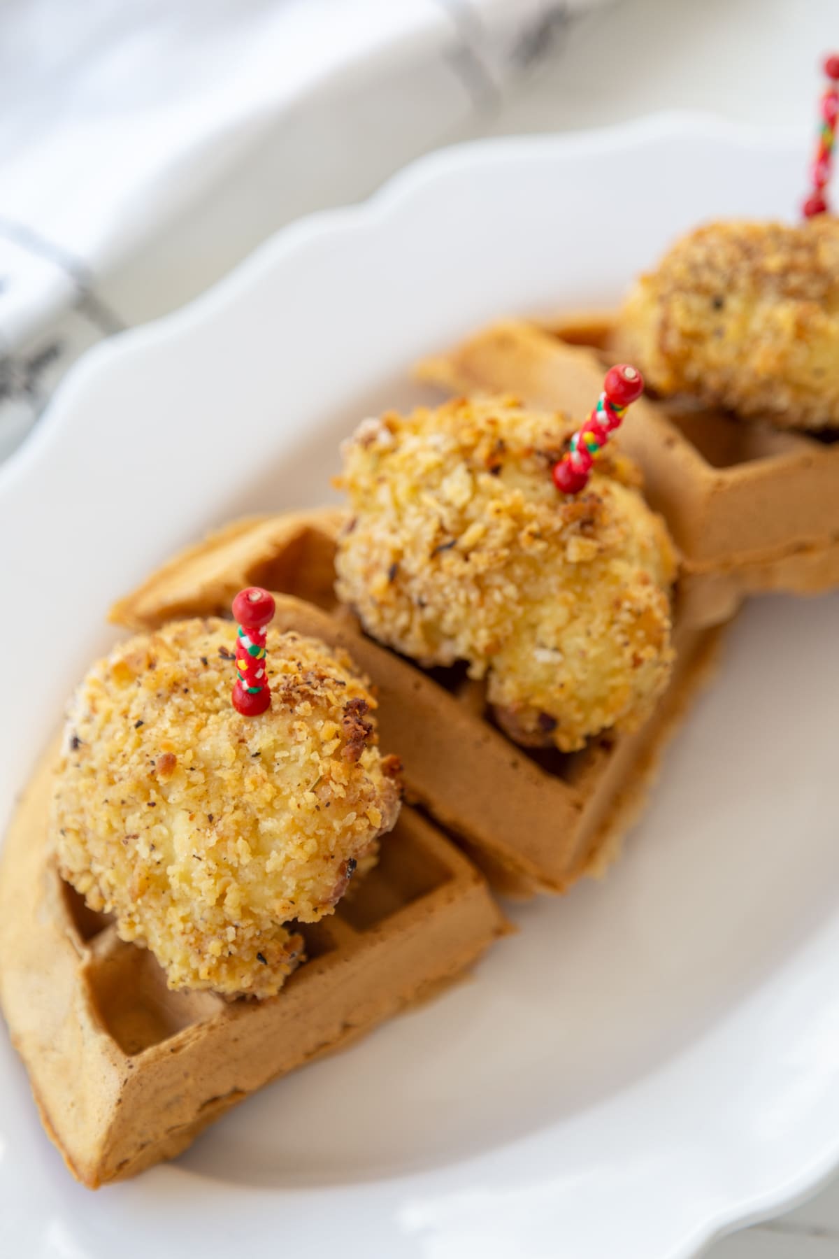 Triangles of belgian waffles on a white platter, topped with crispy cauliflower with a red skewer holding them together.